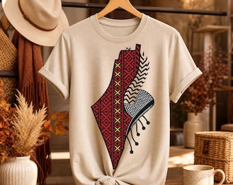 May include: A beige short-sleeved t-shirt featuring a large graphic print of a map shape filled with red and black geometric patterns, yellow cross-stitch details, and a black and white checkered section with dangling circular accents.