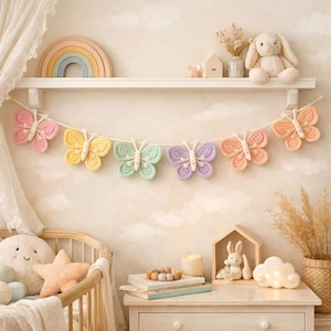 Peut inclure: Une guirlande de papillons au crochet aux couleurs pastel. La guirlande présente des papillons dans les tons rose, jaune, vert, violet et orange. Les papillons sont reliés par un cordon blanc. La guirlande est exposée au-dessus d'un berceau dans une chambre d'enfant.