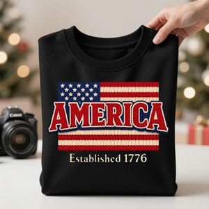 Peut inclure: Sweat-shirt noir à col rond avec un motif tricoté du drapeau américain. Le mot "AMERICA" est en rouge et bleu, et en dessous, le texte "Established 1776".