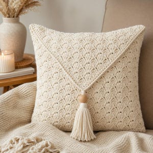 Pode incluir: Almofada de crochê creme com um padrão de concha texturizado e uma aba triangular. Uma conta de madeira e uma borla pendem da ponta da aba. A almofada está sobre um cobertor de malha creme, sugerindo um tema de decoração de casa aconchegante.