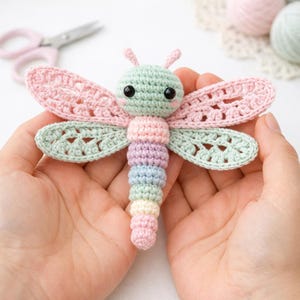 Peut inclure: Une libellule au crochet faite à la main aux couleurs pastel. La libellule a une tête verte, des joues roses et des yeux noirs. Les ailes sont roses et vertes, et le corps est rayé de couleurs arc-en-ciel pastel. La libellule est tenue dans des mains ouvertes.