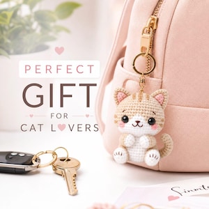 Peut inclure: Un porte-clés chat en crochet fait de fil beige et blanc, avec de grands yeux noirs, des joues roses et un anneau en métal doré. Le porte-clés est attaché à la fermeture éclair d'un sac à dos rose pâle. Le texte indique PERFECT GIFT FOR CAT LOVERS.