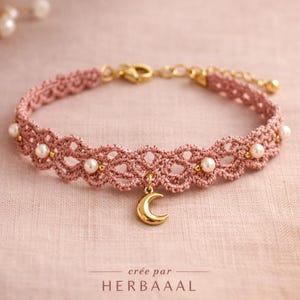 Peut inclure: Un collier ras du cou en crochet rose poudré avec un charm en forme de croissant de lune doré. Le collier est orné de fausses perles et d'accents dorés. Le texte "créé par HERBAAAAL" est en bas.