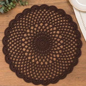 May include: A round, dark brown crocheted doily with a scalloped edge. The openwork design features a central circle and radiating patterns. The doily is placed on a wooden surface, suggesting a decorative or functional use in a home setting.