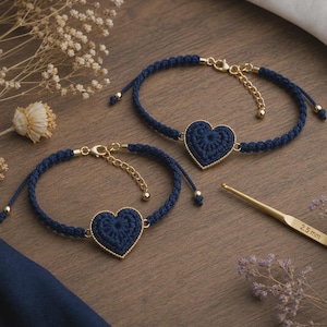 May include: Two navy blue crochet heart bracelets with gold-colored accents. Each bracelet features a heart-shaped charm and adjustable chain. The bracelets are displayed on a wooden surface with dried flowers and a crochet hook.