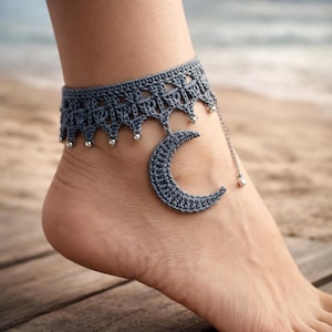 Puede incluir: Una tobillera de ganchillo gris con un dije de media luna y pequeñas cuentas plateadas. La tobillera se usa en un tobillo, con una superficie de madera en primer plano y una playa en el fondo. Un accesorio hecho a mano.