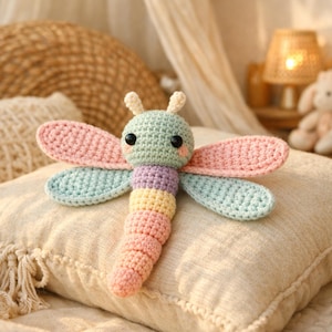 Peut inclure: Une libellule au crochet faite à la main dans des couleurs pastel. La libellule a une tête vert clair, des ailes roses et bleues, et un corps segmenté dans des tons de violet, jaune et rose. Elle repose sur un coussin beige.