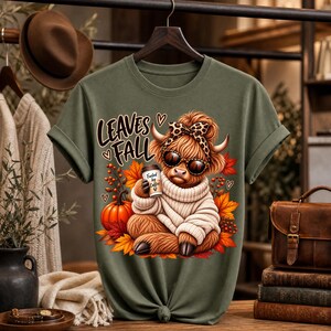 Peut inclure: Un t-shirt vert olive avec une illustration d'une vache highland poilue portant un nœud léopard et des lunettes de soleil, tenant une tasse de café. Le texte indique LEAVES FALL. La vache est entourée de citrouilles et de feuilles d'automne.