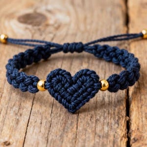 May include: Navy blue macrame bracelet featuring a heart-shaped design. The bracelet has gold-colored beads and an adjustable sliding knot closure. The bracelet is made of woven cord and is displayed on a wooden surface.