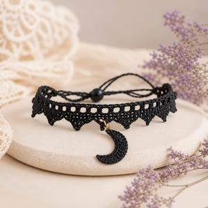 May include: A black crochet choker necklace with a crescent moon charm. The necklace features a scalloped edge design and a small black bead for closure. The moon charm is gold-colored. The necklace is displayed on a neutral-colored surface.