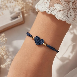 May include: A navy blue crochet bracelet with a heart-shaped charm and gold-colored beads. The bracelet is worn on a person's arm, with a white lace sleeve visible in the background. The bracelet is a delicate piece of jewelry.