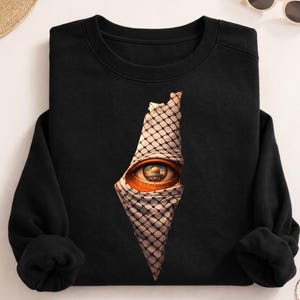 Peut inclure: Sweat-shirt noir avec un motif graphique. Le motif représente un œil avec un élément architectural du Moyen-Orient dans l'iris, superposé à un contour de carte. L'œil est partiellement recouvert d'un tissu à motifs.
