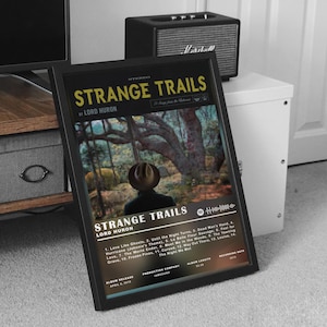 Può includere: Un poster incorniciato nero con un titolo giallo e bianco "Strange Trails" di Lord Huron. Il poster presenta una foto di una persona con un cappello in piedi in un bosco. La tracklist dell'album è elencata sotto il titolo.