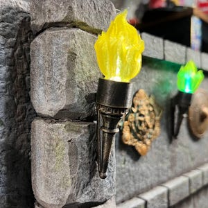 May include: A close-up of a stone wall with two decorative torches and a wall sconce. The torches have a yellow and green flame effect. The wall sconce is bronze-colored.