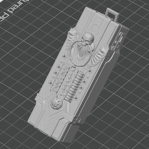 May include: A gray 3D printed rectangular case featuring a skull motif with a ring through its mouth, intricate mechanical detailing, and a small loop at the top. The surface displays a complex relief pattern with various knobs and ridges.