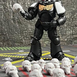 Può includere: Un action figure di space marine in armatura nera con spallacci bianchi e un emblema dorato sul petto, che tiene in mano un piccolo teschio bianco. Diversi accessori per elmi a forma di teschio bianco non dipinti sono sparsi sul terreno.