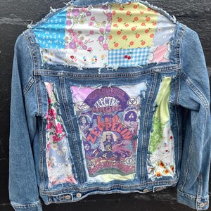 May include: A light blue denim jacket with patchwork fabric accents. The back features a vintage concert poster for Electric Magic featuring Led Zeppelin. Floral and patterned fabrics in various colors are sewn onto the jacket.