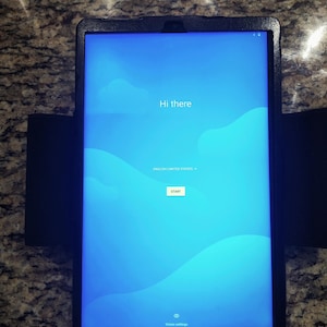 May include: A tablet device in a black protective case displaying a blue screen with the text Hi there, English (United States), START, and Vision settings. The screen features a light blue abstract wave pattern across the background.
