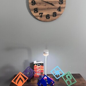 May include: A wooden clock with black numbers hangs above a table displaying colorful geometric art pieces. The art pieces are in orange, purple, blue, light blue, and green. A book titled "Unzed" is also on the table.