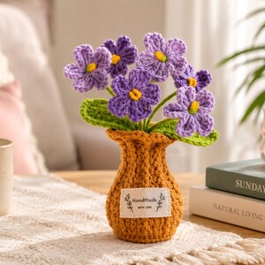 Peut inclure: Un bouquet crocheté de fleurs violettes avec des centres jaunes et des feuilles vertes disposé dans un vase texturé jaune moutarde. Une étiquette en tissu rectangulaire blanche sur le devant du vase porte le texte Handmade with love en noir.