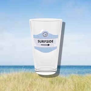 Puede incluir: Un vaso de pinta transparente con una etiqueta azul claro y blanca que dice "SURFSIDE" con una flecha que apunta hacia la derecha. El fondo muestra una escena de playa con cielo azul y océano.