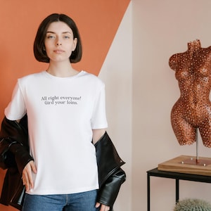 Puede incluir: Una camiseta blanca con el texto "All right everyone! Gird your loins" en negro. La persona lleva una chaqueta de cuero negra y vaqueros azules. Una escultura decorativa de torso color cobre está sobre una mesa negra.