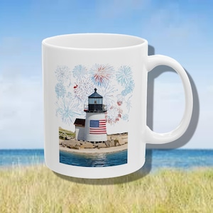 May include: White ceramic mug featuring a lighthouse with an American flag design and fireworks. The lighthouse is set on a sandy beach with blue water and sky in the background. The mug has a curved handle.