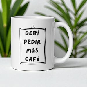 Puede incluir: Taza de café de cerámica blanca con un diseño enmarcado en negro. El diseño presenta las palabras "DEBI PEDIR MÁS CAFÉ" en una fuente de estilo manuscrito. La taza tiene un asa curva y está sobre un fondo de follaje verde.