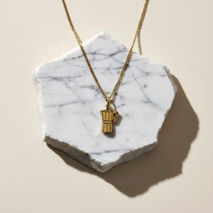 May include: A gold-coloured metal necklace featuring a small pendant shaped like a moka pot coffee maker. The pendant hangs from a delicate chain, showcasing a polished finish with engraved lines that define the classic octagonal coffee pot form.