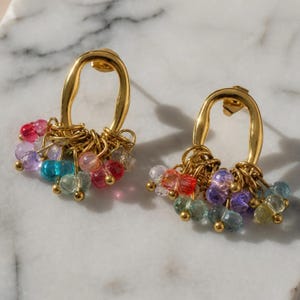Puede incluir: Un par de pendientes dorados con un marco ovalado de superficie texturizada. Cada pendiente está adornado con un racimo de pequeñas cuentas facetadas multicolores en tonos rosa, morado, azul y verde, suspendidas desde la base.