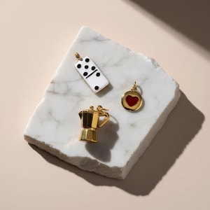May include: Three gold-toned metal charms including a white rectangular domino piece with black dots, a circular charm with a red heart centre, and a three-dimensional moka pot coffee maker, each featuring a small loop for attachment to jewellery.