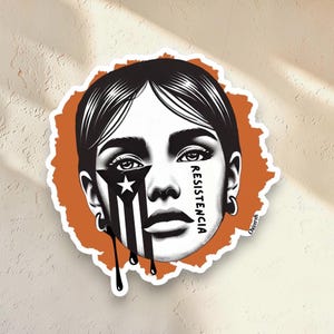 Puede incluir: Una pegatina con una ilustración estilizada del rostro de una mujer con cabello negro y un diseño de la bandera de Puerto Rico en blanco y negro en un lado. La palabra "RESISTENCIA" está escrita verticalmente en la cara. La pegatina tiene un borde naranja.