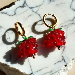 May include: A pair of gold hoop earrings featuring dangling red raspberry charms. Each charm is crafted with translucent red beads clustered to resemble a berry, accented with small green leaves at the top and tiny gold beads at the base.