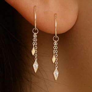 May include: A pair of gold-colored hoop earrings. Each earring features a delicate chain with a small, diamond-shaped charm and a smaller, teardrop-shaped charm. The earrings are worn on a person's ear.