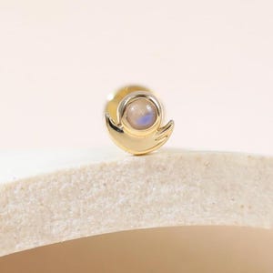 May include: A small, gold-colored stud earring with a round, iridescent stone at the center. The earring has a stylized, curved design. The background is a soft, neutral color, highlighting the jewelry.