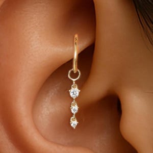 May include: A gold-colored earring with three dangling clear gemstones. The earring is a hoop style, designed to be worn in a pierced ear. The gemstones are round and appear to be set in a delicate, minimalist design.