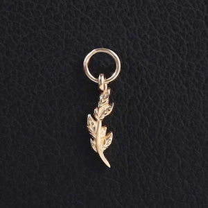 May include: A small gold-toned metal charm shaped like a delicate feather, featuring intricate etched details along its surface. The feather charm is attached to a simple, circular gold-toned jump ring, designed for easy attachment to a chain or bracelet.