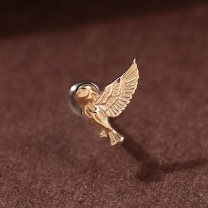 Pode incluir: Um brinco dourado em forma de pássaro com asas abertas. O brinco tem uma pequena cabeça redonda prateada e um design de asa detalhado. O brinco está sobre um fundo castanho.