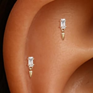 Puede incluir: Dos pendientes dorados con una gema rectangular y transparente y un pequeño dije dorado puntiagudo. Los pendientes se muestran en la oreja de una persona, mostrando el diseño y cómo se ven cuando se usan.
