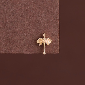 May include: A small gold-toned stud earring shaped like a dragon with outstretched wings and a long tail. The piece features a metallic finish and is positioned at the edge of a textured brown surface, showcasing its detailed, symmetrical design.