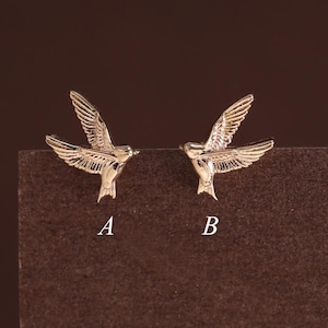 May include: A pair of gold-toned metal bird stud earrings, each shaped like a bird in flight with detailed wing feathers. The earring on the left is labeled with the letter A, and the earring on the right is labeled with the letter B.