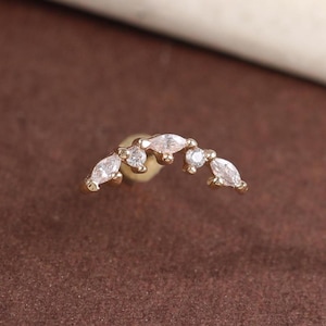 May include: A gold-colored earring with a curved design. The earring features several clear, marquise-cut gemstones arranged in an arc. The earring is set against a brown background.