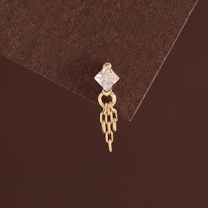 May include: A gold-toned stud earring featuring a clear, square-cut gemstone at the top. Attached to the base of the gemstone is a small, dangling gold-toned chain element consisting of three short, interconnected vertical links that hang downward.
