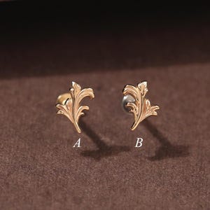 Op de afbeelding: Twee goudkleurige oorstekers in de vorm van een blad. Elke oorbel heeft een gedetailleerd, sierlijk ontwerp met delicate rondingen en texturen. De oorbellen worden gepresenteerd op een bruin oppervlak, met de letters "A" en "B".