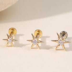 May include: Three gold-toned stud earrings, each featuring a six-pointed star design with a small, clear, round-cut gemstone set in the center. The earrings have a flat, circular backing post and a polished metallic finish, arranged in a horizontal row.