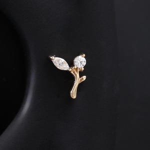 May include: A gold-colored stud earring featuring a delicate branch design. The earring has two clear, sparkling gemstones: one marquise-cut and one round-cut. The earring is set against a dark background.