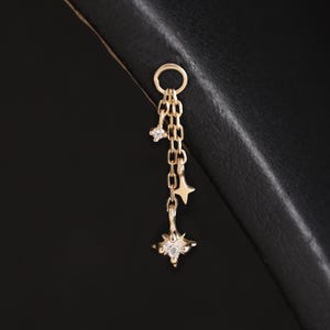 May include: A gold-toned earring charm featuring a small circular hoop connected to two delicate chains of varying lengths. The chains are adorned with three small star-shaped pendants, each set with a tiny clear stone at its center point.