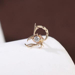 May include: A gold-colored scorpion-shaped earring with a small, clear gemstone at its center. The scorpion's tail curves upwards, and its claws are open. The earring is set against a white surface with a brown background.
