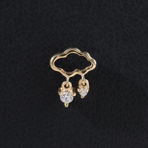 Puede incluir: Un charm en forma de nube dorada con dos colgantes en forma de lágrima. Cada colgante presenta una gema transparente de talla redonda. El charm está sobre un fondo negro.