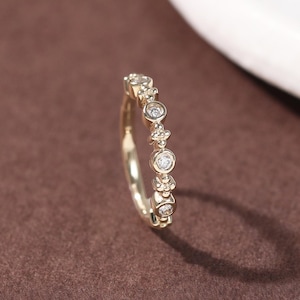 Può includere: Un delicato anello d'oro con una serie di piccole pietre preziose trasparenti. L'anello ha una fascia sottile e le pietre preziose sono incastonate in un motivo ripetuto. L'anello è esposto su una superficie marrone con uno sfondo bianco.